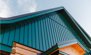 Close-up view of green vertical metal cladding on the gable end of a modern structure, featuring clean architectural lines and exposed wood details under a clear blue sky