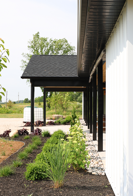 Side view of a modern home clad in white vertical metal siding with black soffits and trim, landscaped with shrubs and a concrete walkway leading to the entrance