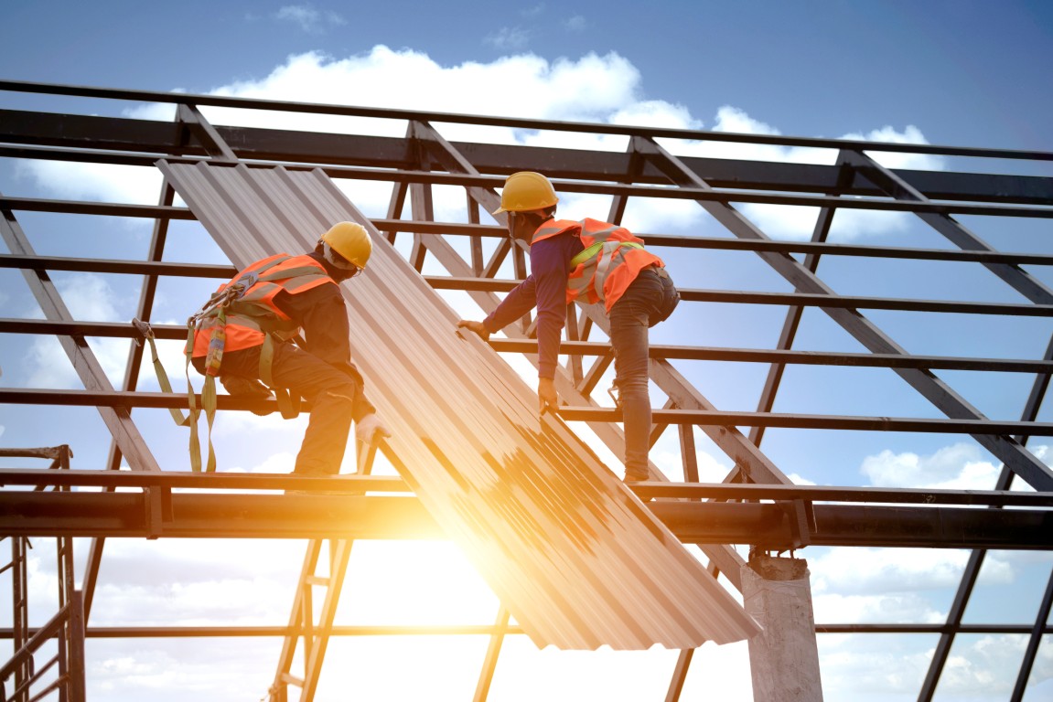 Two construction workers in safety gear installing corrugated metal roofing panels on a steel frame under bright sky