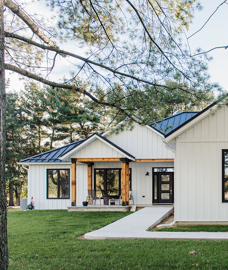 Stylish modern farmhouse with white vertical board and batten siding, black standing seam metal roof, natural wood posts, and large black-trimmed windows nestled among mature pine trees