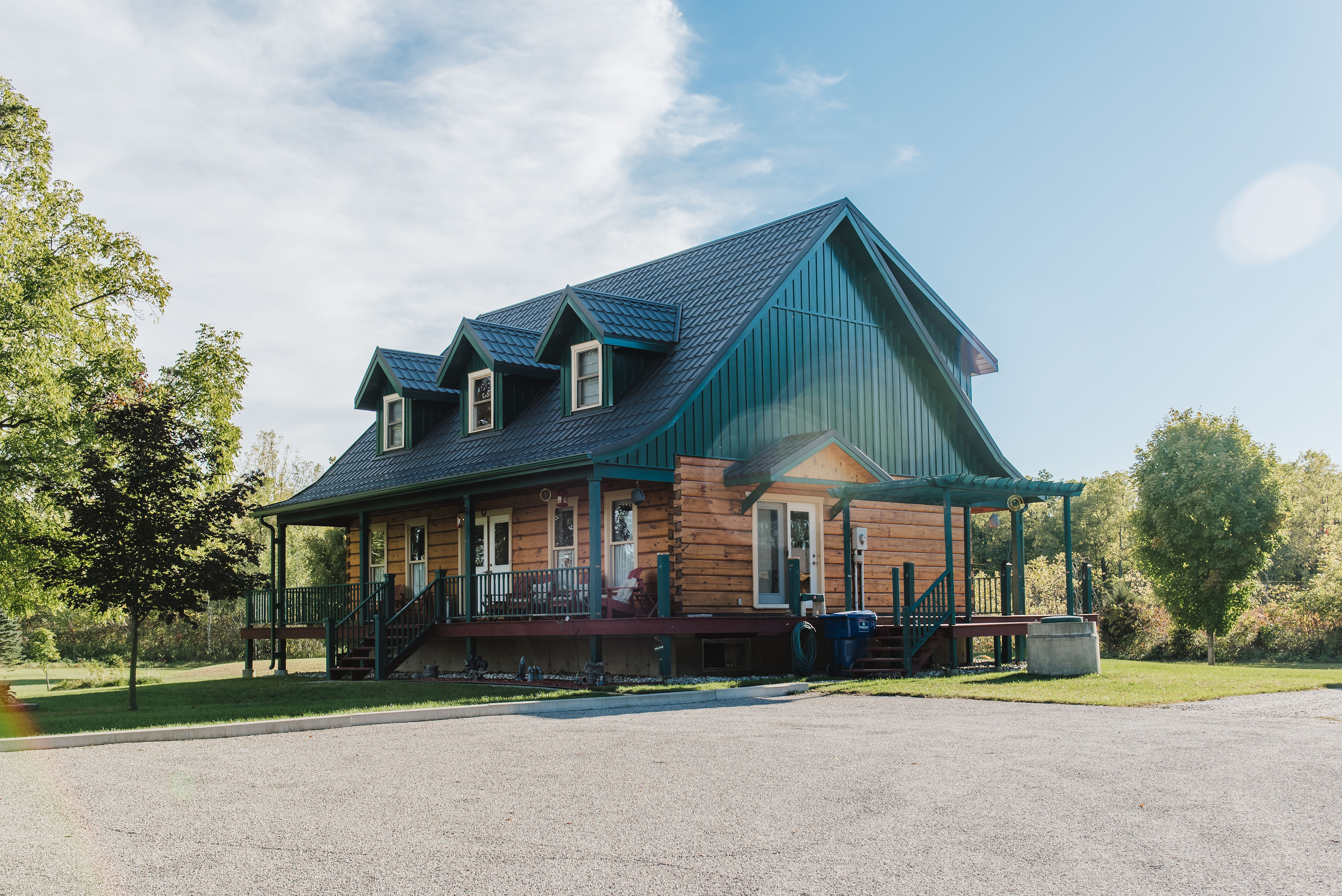 Charming log-style cabin with green standing seam metal roofing, wood siding, and front dormers, set in a lush rural landscape under a clear blue sky